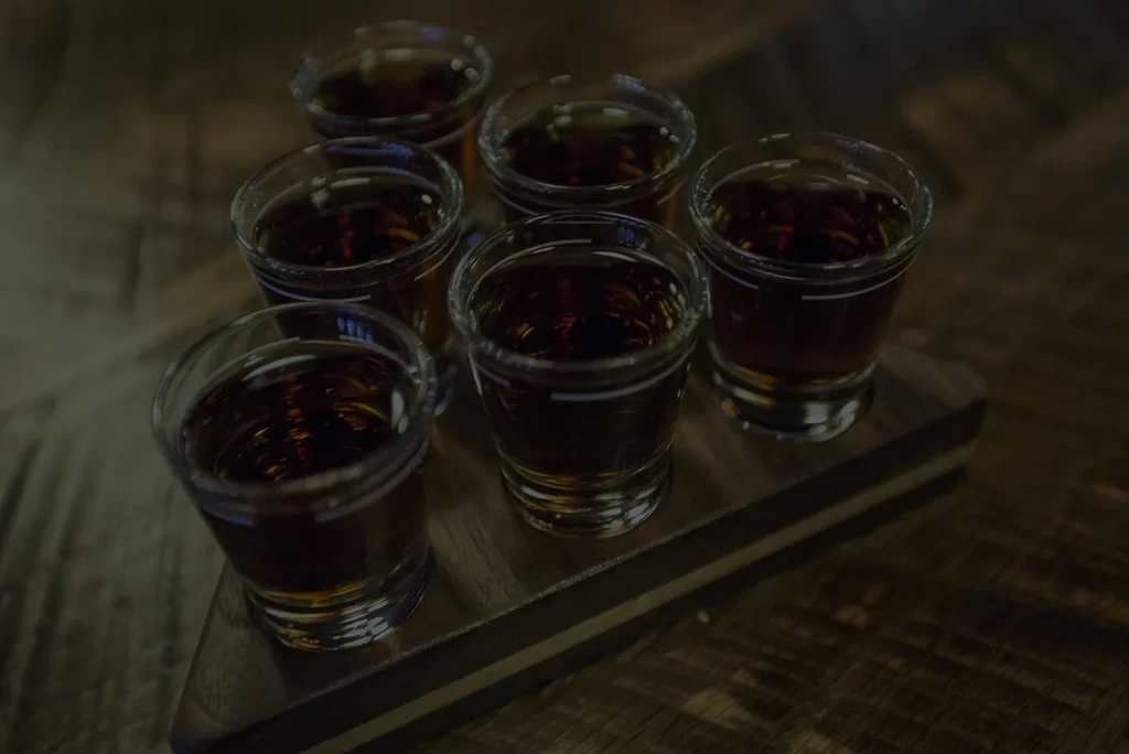 Six shot glasses filled with dark amber liquid on a rustic board.