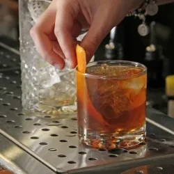 Hand garnishing an amber cocktail with an orange peel.