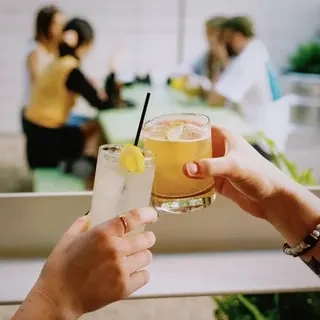 Two hands clinking cocktail glasses, surrounded by blurred people.