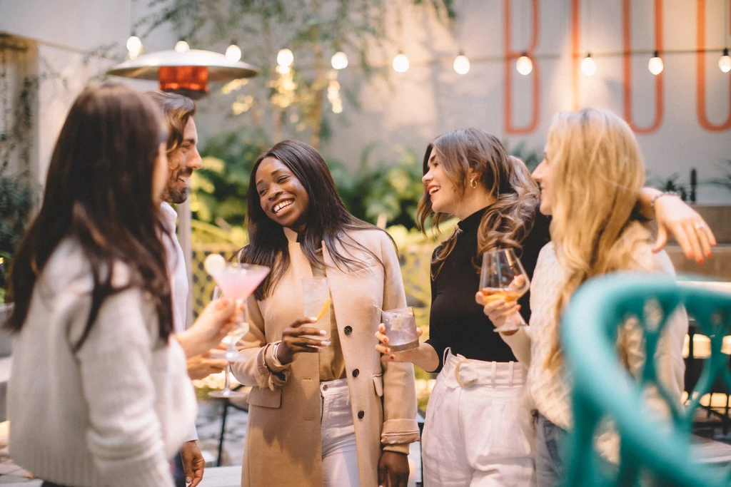 Five people with drinks socialize at an outdoor bar at night.