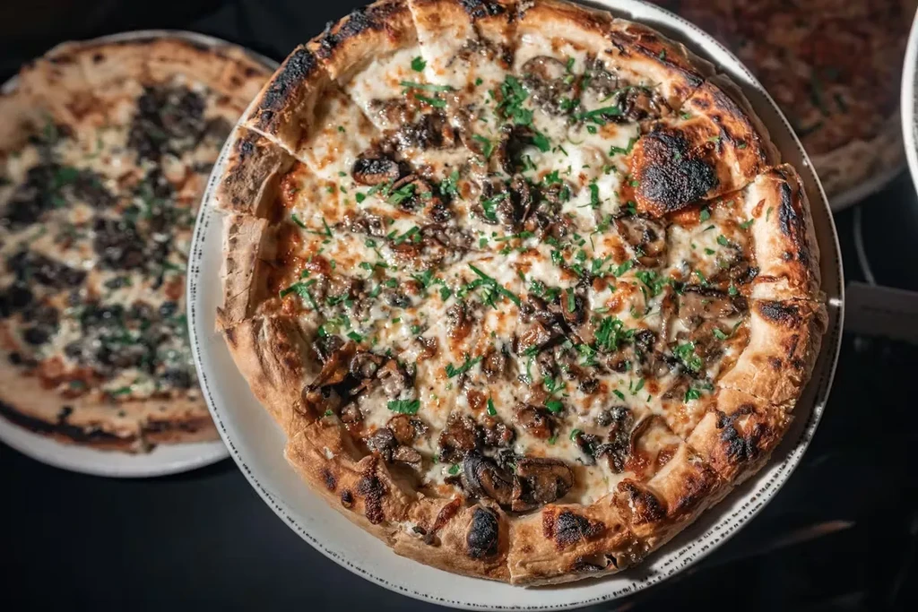 Two pizzas, one in focus, topped with cheese, mushrooms, and herbs.