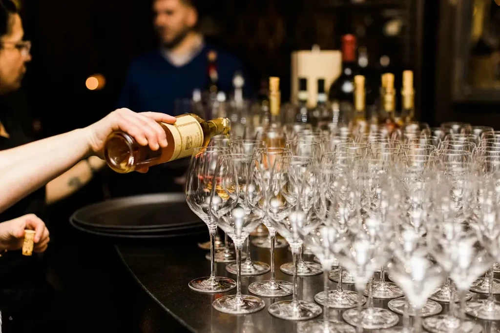 Hands pour amber liquid into a wine glass; many empty glasses lined up.