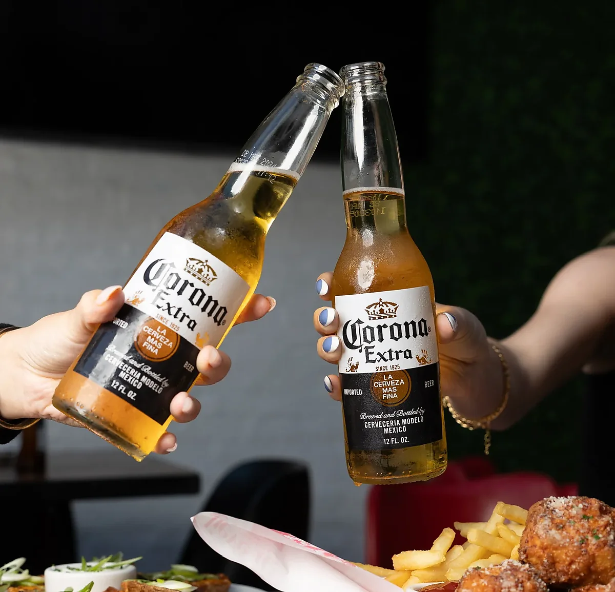 Two Corona bottles toasting above plates of fries and wings