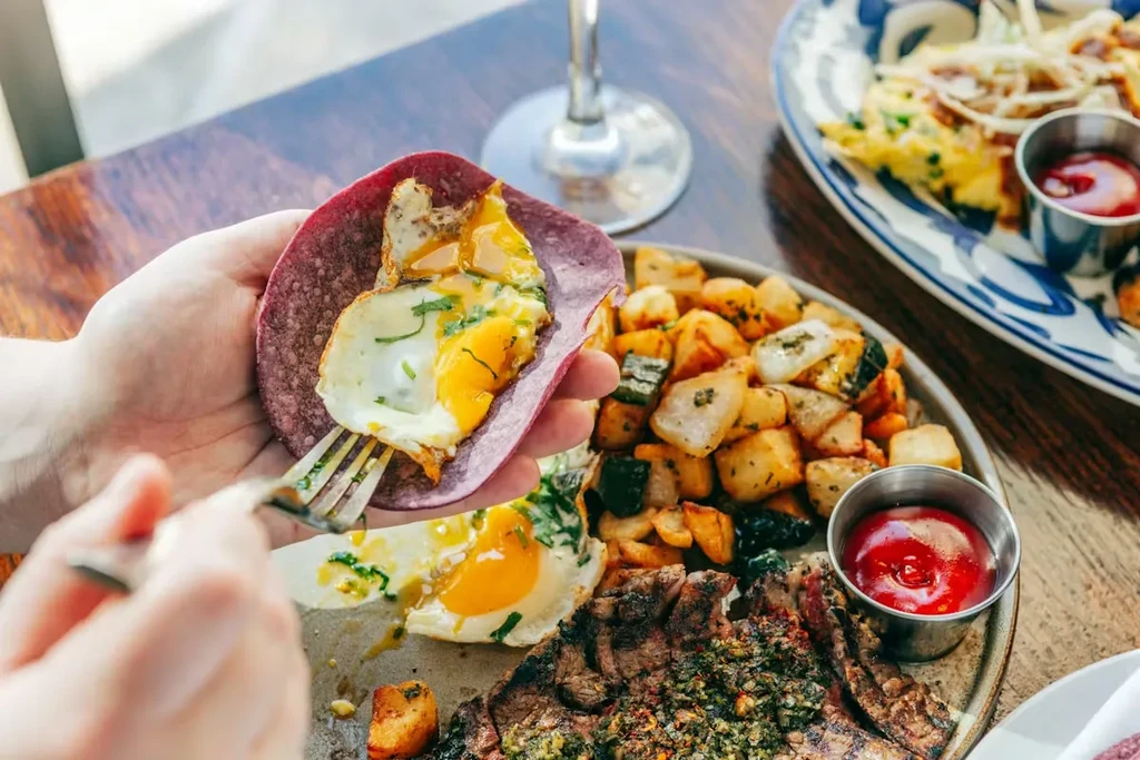 Hand holding a purple tortilla with fried egg on a breakfast spread.