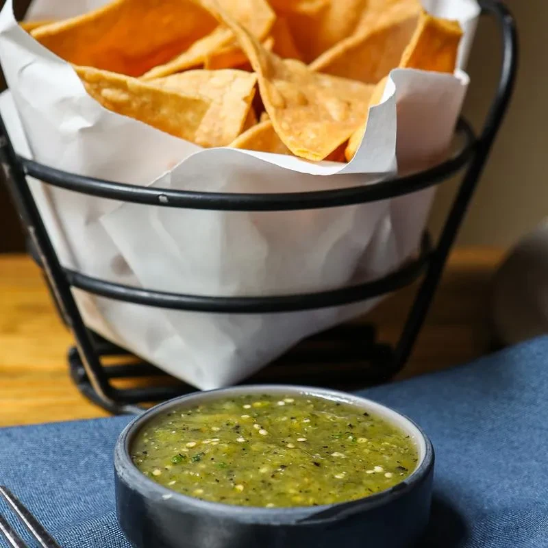 Tortilla chips in a basket with a bowl of green salsa.