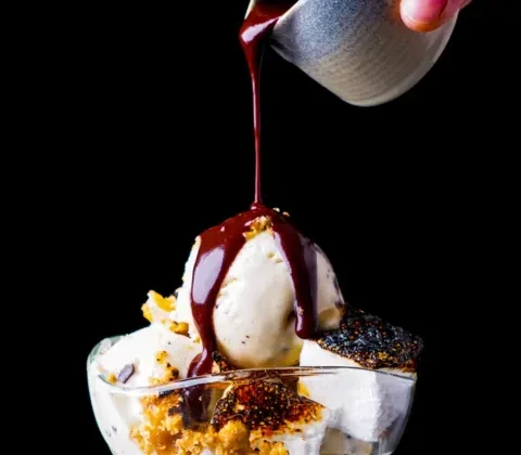 Vanilla ice cream with chocolate sauce being poured onto it.