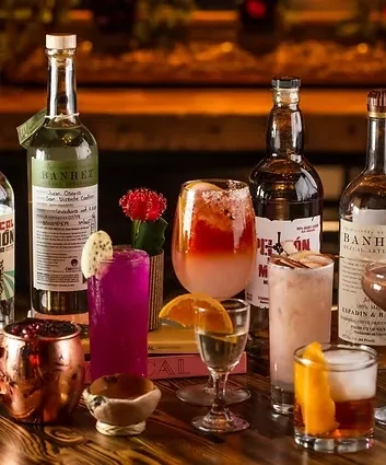 Cocktails and spirits, including Banhez mezcal, on a wooden surface.
