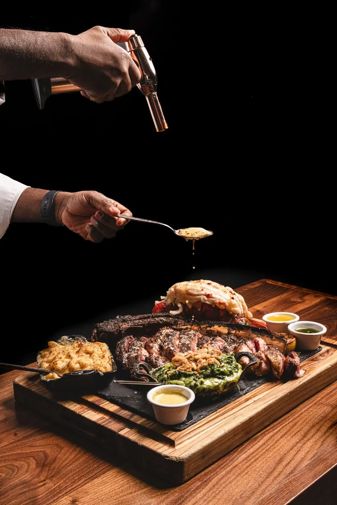 Hands torching and drizzling sauce on tomahawk steak and lobster meal.