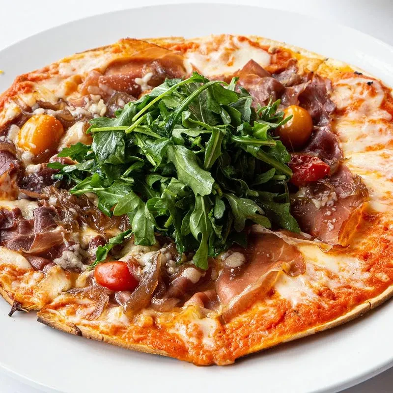 Pizza or flatbread with prosciutto, arugula, cherry tomatoes, and caramelized onions.