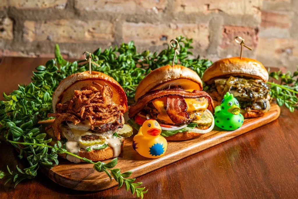 Three gourmet hamburgers on a wooden board with two rubber ducks.