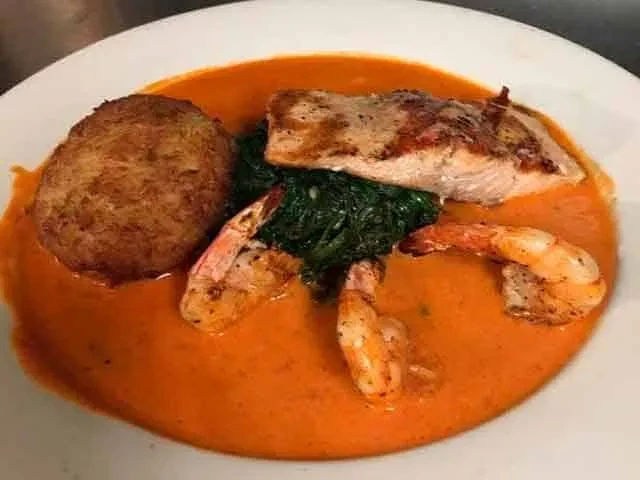 Plated pan-seared fish, shrimp, vegetables, sauce, and a croquette.