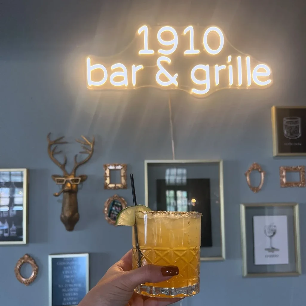 Hand holding a lime-garnished cocktail with "1910 bar & grille" neon sign.