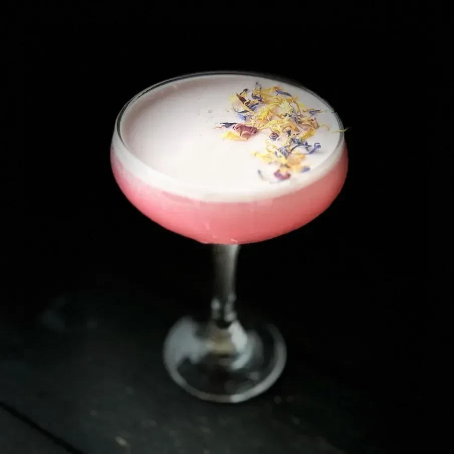 Pink cocktail in coupe glass with white foam and colorful flower petals.
