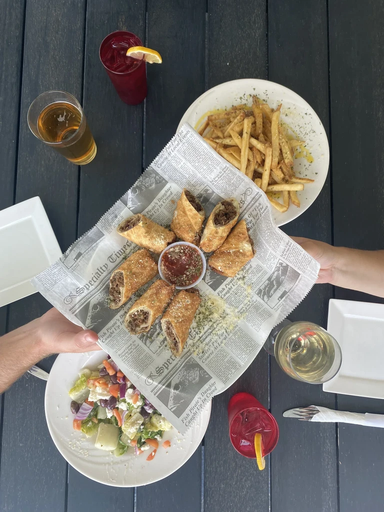 Overhead view of hands sharing crispy rolls, fries, salad, and drinks.