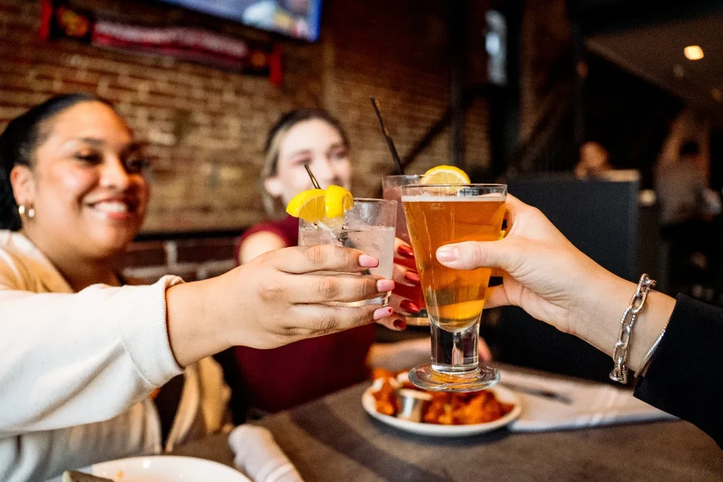 Three hands toast cocktails and beer; blurred smiling women behind.