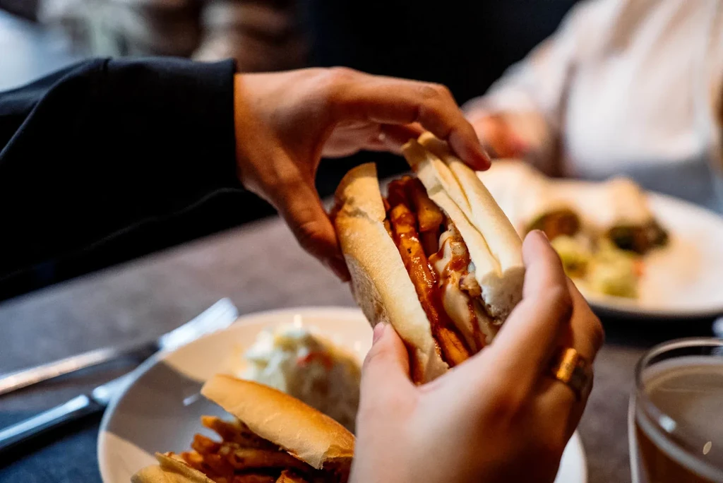 Hands holding a large sandwich filled with fries, red sauce, and meat.