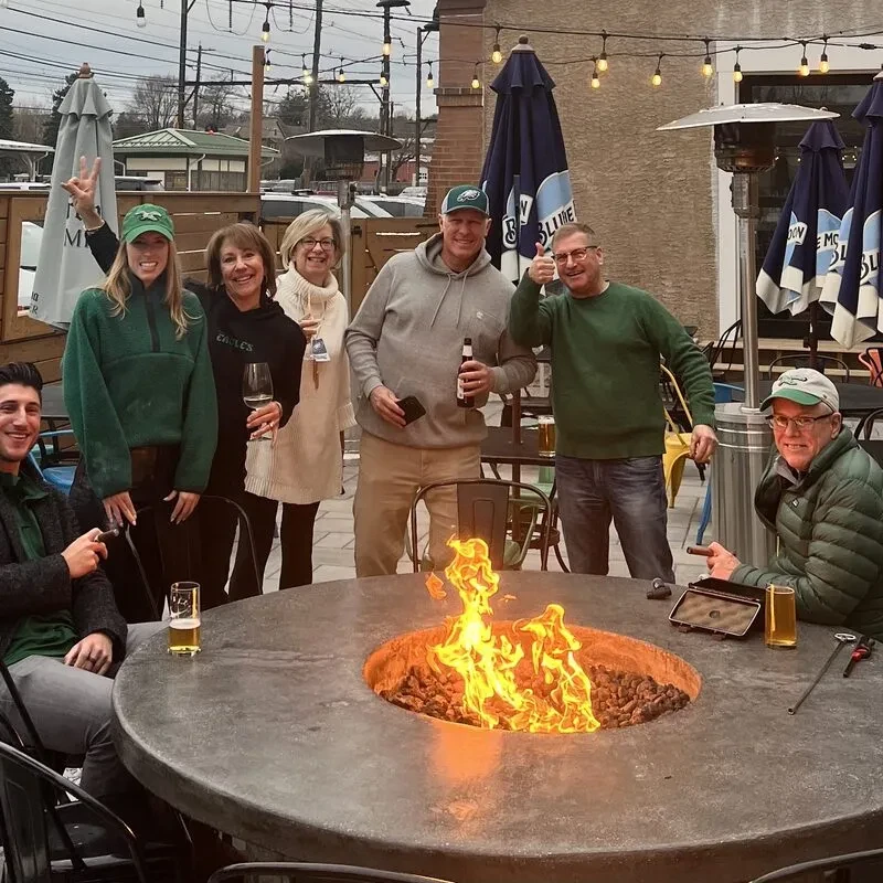 Seven people gather around a burning fire pit on an outdoor patio.