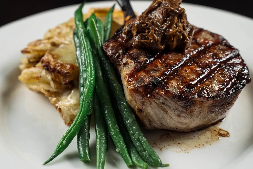 Plated grilled steak, green beans, and au gratin potatoes with dark sauce.