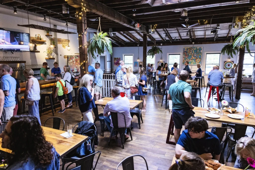 Bustling restaurant interior with wooden beams, plants, abstract art, and many patrons.