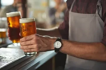 Bartender's hands place two glasses of amber beer on a metal bar.