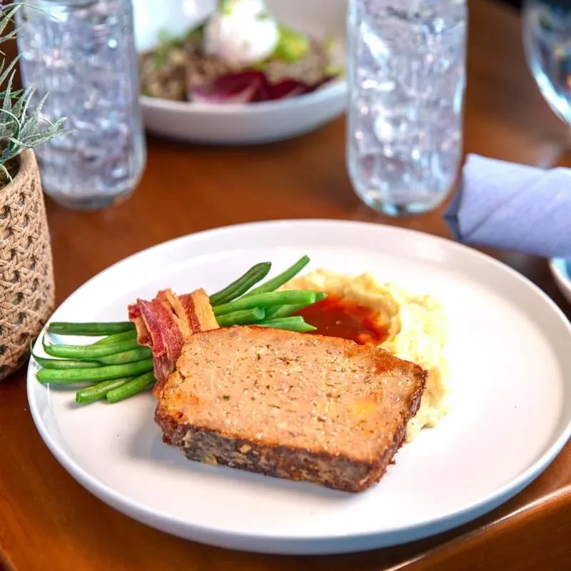 Meatloaf, bacon-wrapped green beans, and mashed potatoes on a white plate.