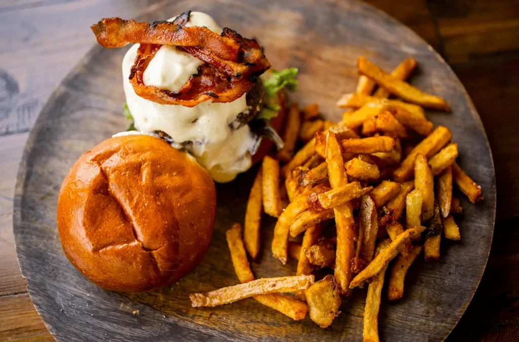 Disassembled gourmet burger, bacon, cheese, lettuce, fries, on wooden board.