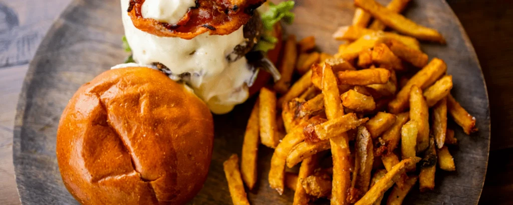 Gourmet burger with creamy sauce, bacon, and fries on a wooden board.