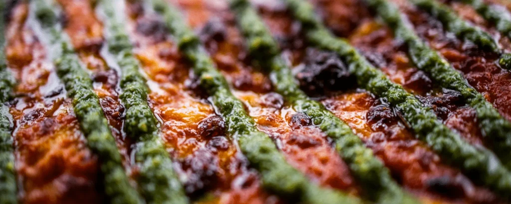 Close-up of baked food: reddish-orange base, green pesto lines, caramelized texture.