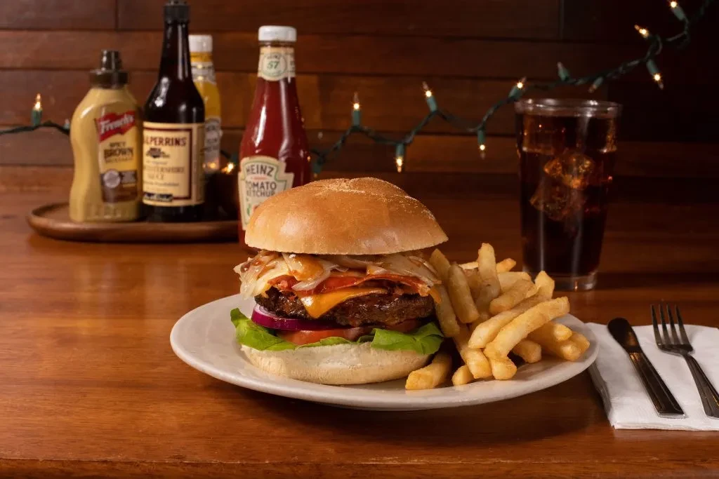 Loaded burger and fries, drink, condiments, string lights on wooden table.