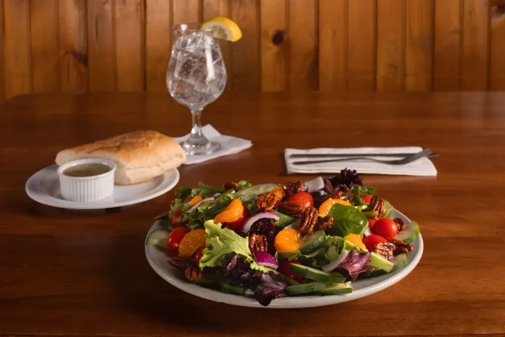 Vibrant salad, bread, sauce, water, and cutlery on a wooden table.