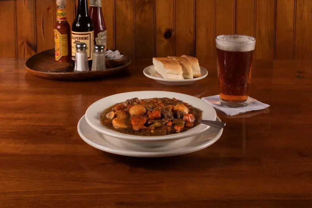 Hearty stew, bread, beer, and condiments on a rustic wooden table.