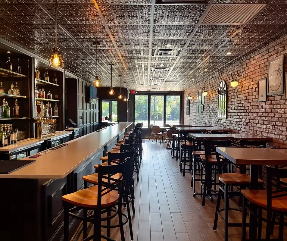 Well-lit restaurant interior with long bar, high-top tables, and brick wall.