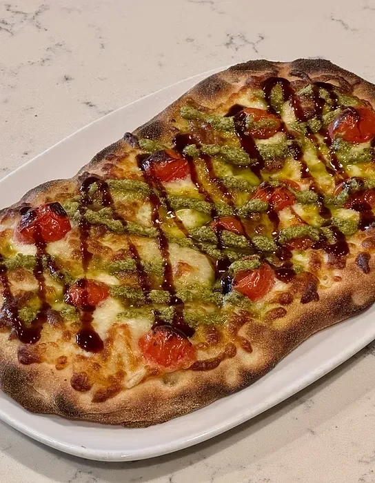 Rectangular flatbread pizza with cheese, tomatoes, pesto, and balsamic glaze.