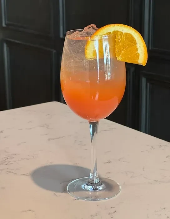 Orange and pink layered drink, ice, orange slice in a wine glass.