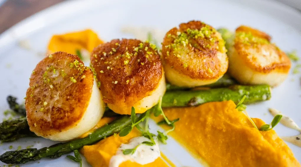 Seared scallops, asparagus, orange puree, and green garnish on white dish.