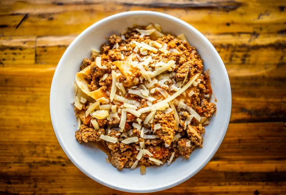 Flat pasta with ground meat sauce and shredded cheese in white bowl.