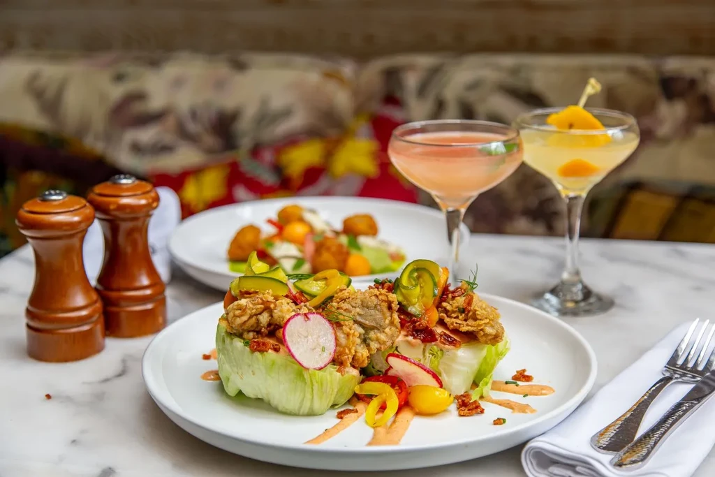 Plated wedge salad, cocktails, and other food on a white marble table.