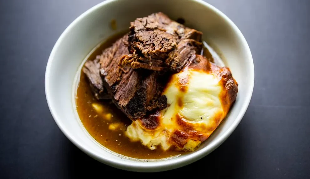 Overhead shot of braised meat and cheesy potato gratin in a white bowl.