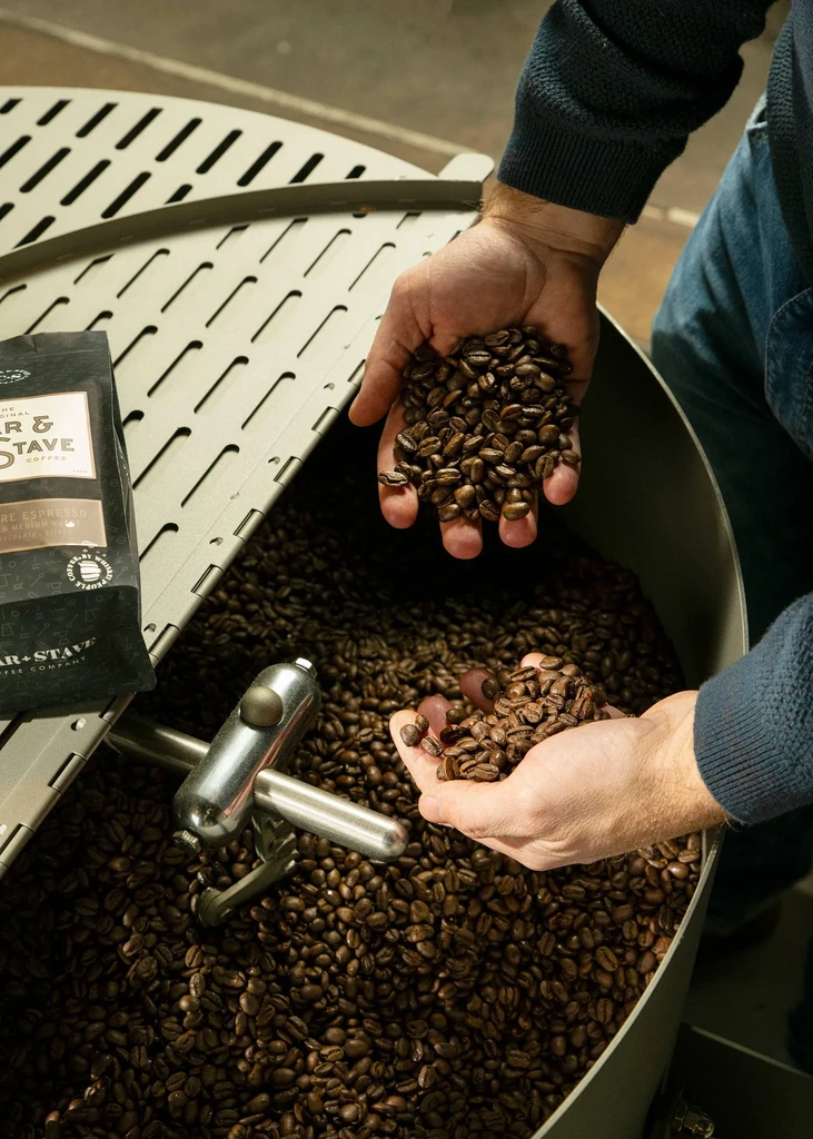 Hands hold fresh, dark roasted coffee beans in a large roaster.