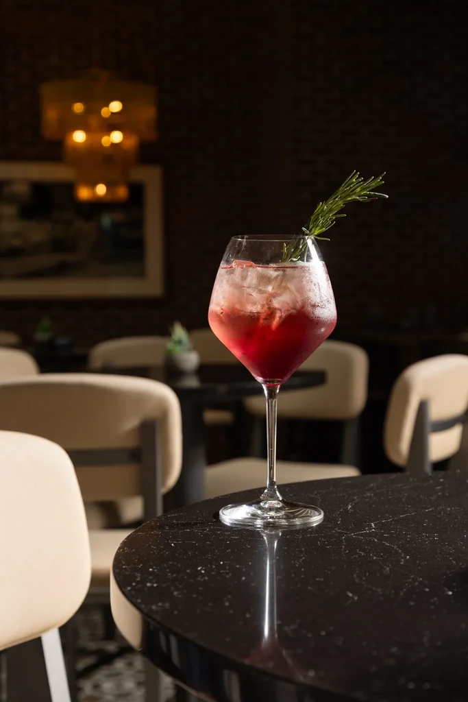 Reddish-pink cocktail with ice, rosemary, in stemmed glass on marble.