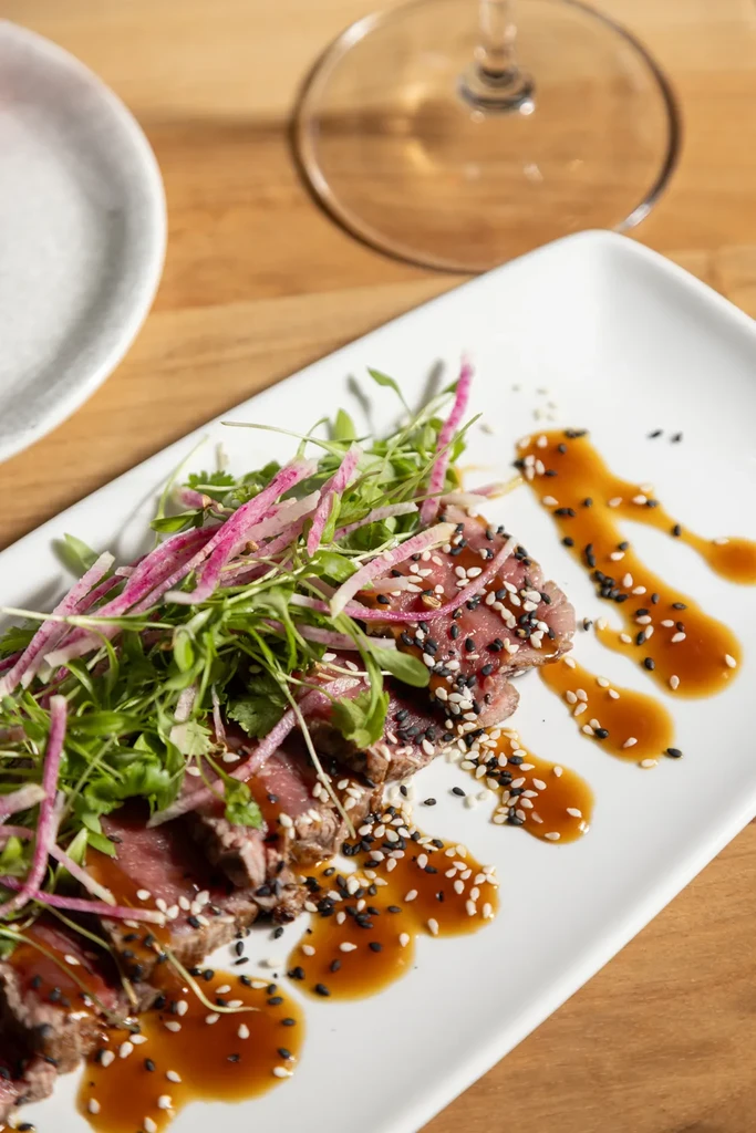 Seared reddish-pink meat with sesame, microgreens, radish, and amber sauce.
