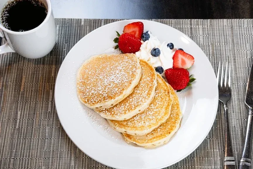 Overhead: Pancakes, berries, whipped cream, coffee, and utensils on placemat.