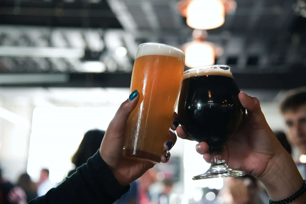 Two hands toast beer glasses: light lager, dark stout, blurred bar background.