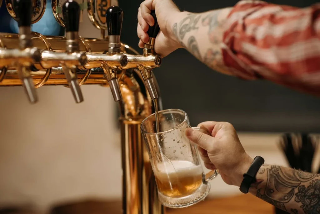 Tattooed hands pour foamy amber beer from a golden draft tap.