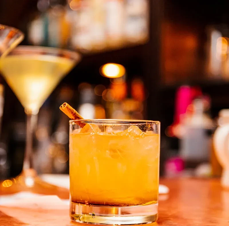 Close-up of amber drink with cinnamon stick, martini glass in blurred bar.