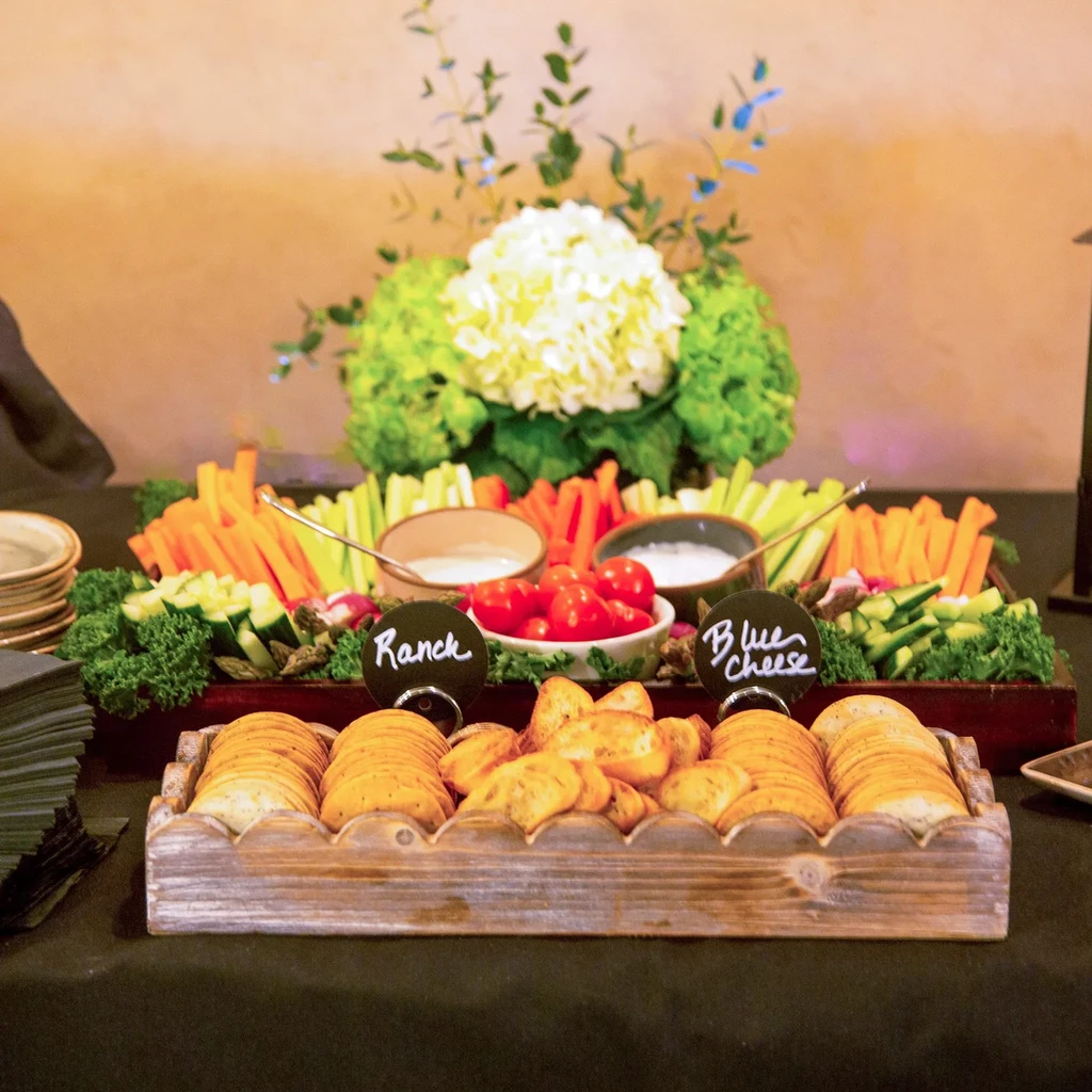 Rustic wooden tray of crudités, dips, cherry tomatoes, and crackers.