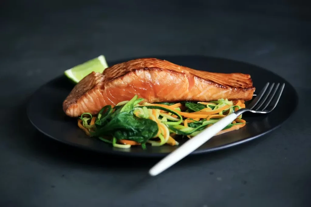 Crispy salmon, colorful spiralized vegetables, and lime on a dark plate.
