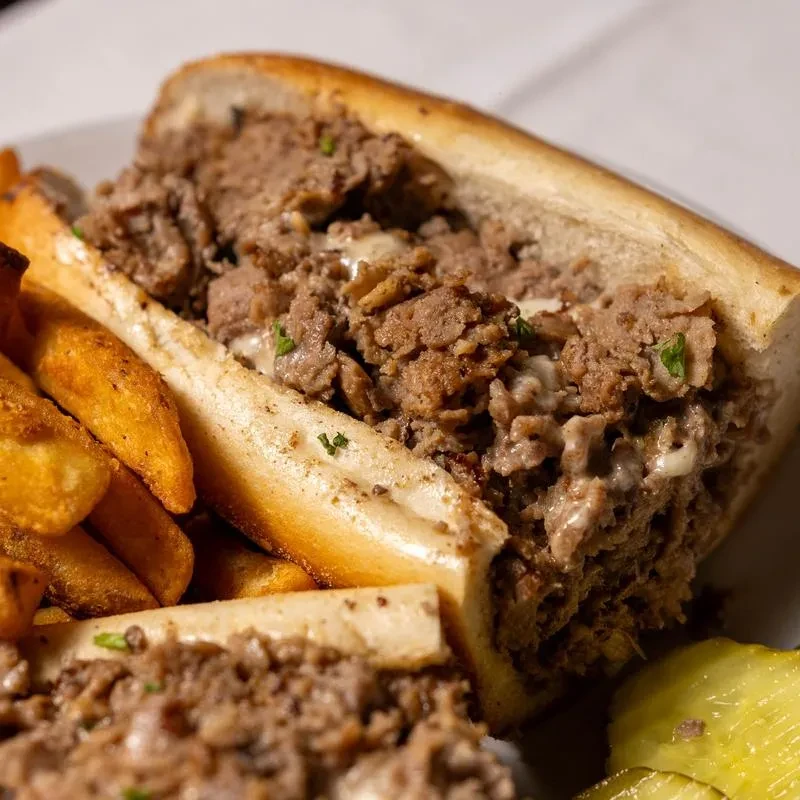 Cheesesteak sandwich, golden fries, and dill pickles on a white plate.