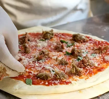 Gloved hand arranging uncooked pizza with sauce, meat, cheese, and basil.
