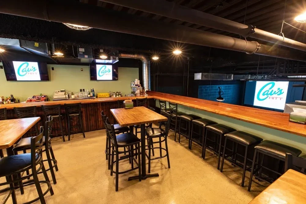 Restaurant bar with high-top tables, industrial ceiling, and Cavi's PITT TVs.